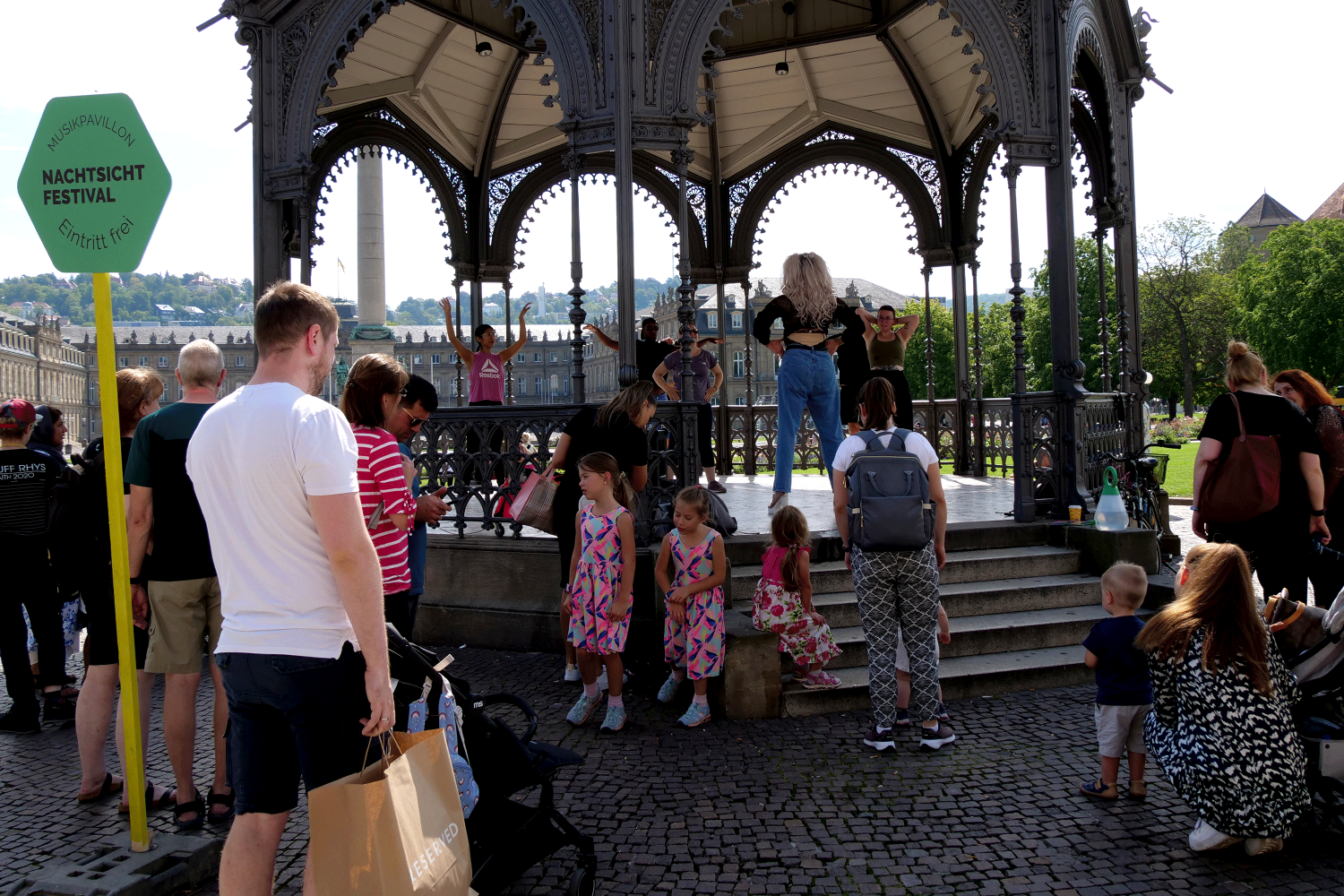 Zu sehen ist der Musikpavillion in der Innenstadt Stuttgart. Um den Pavillion herum laufen Passant*innen. Im Pavillion ist die Drag Queen Dua Look zu sehen, die eine Queerobic Stunde anleitet.