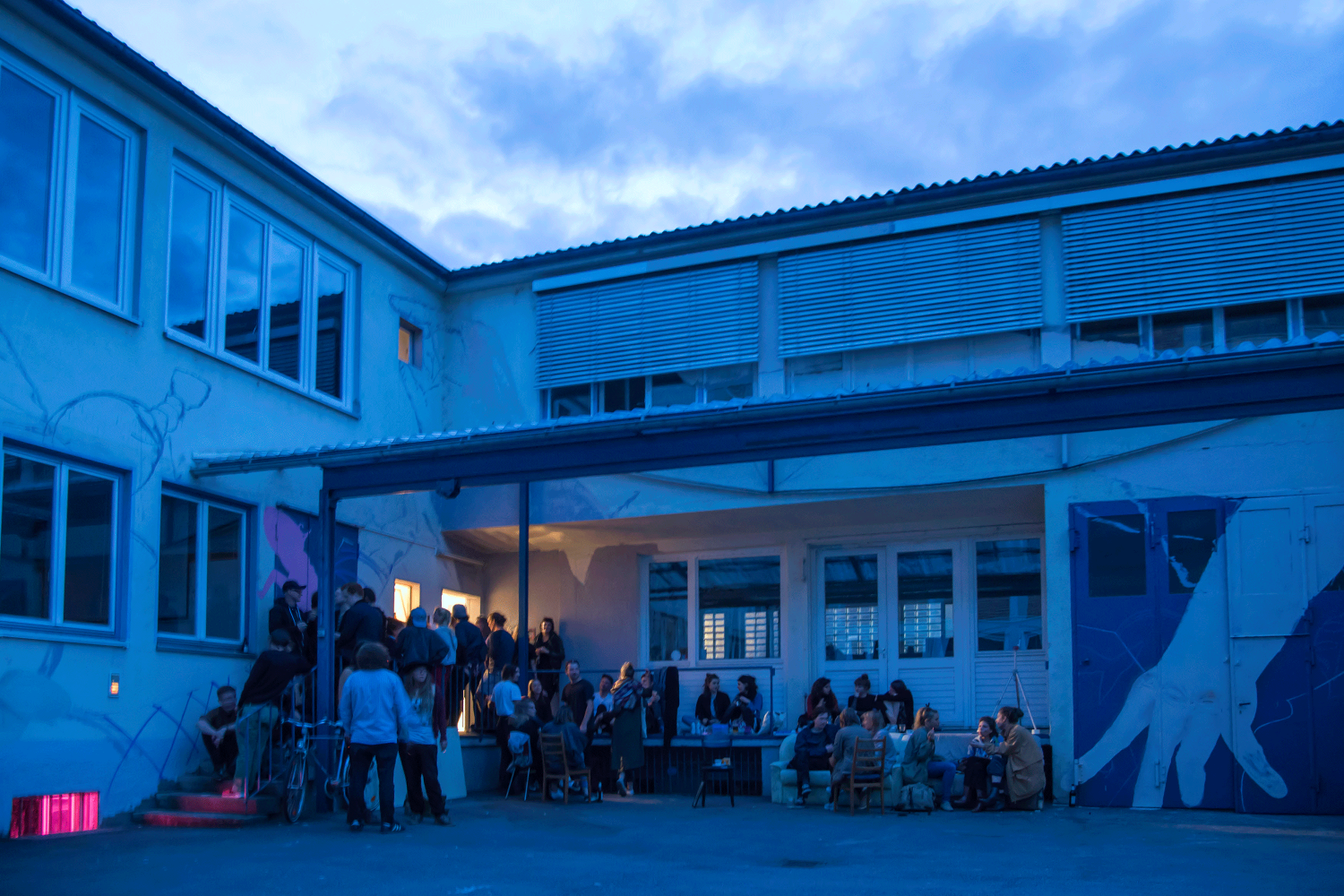 Eine Masse Menschen versammelt sich auf der Veranda vor der Schmuckmanufaktur. Es dämmert. Aus einem Kellerzimmerfenster strahlt pinkes Licht. Die Menschen trinken und unterhalten sich.