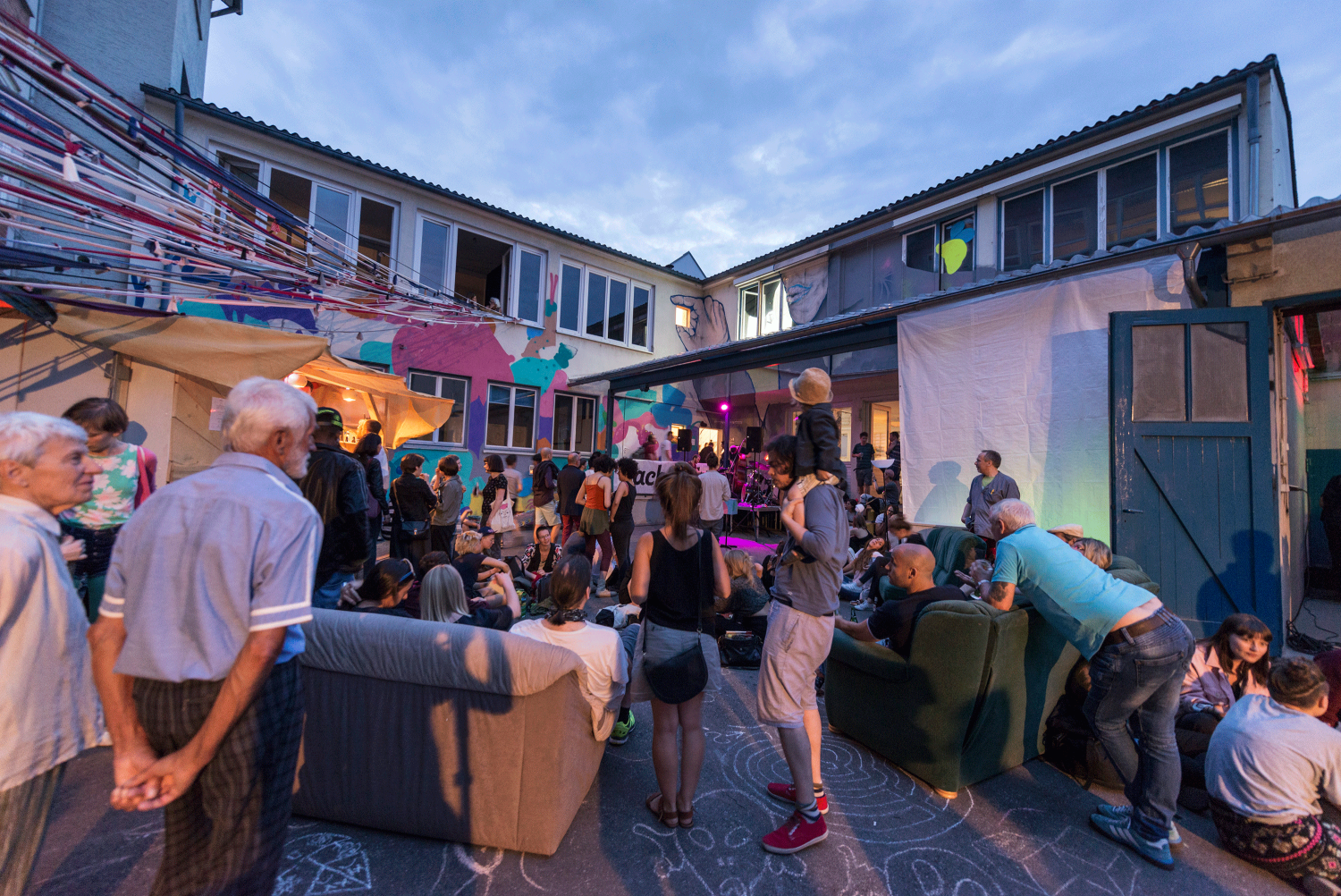 Das Foto zeigt den Innhof der Manufaktur am frühen Abend, die Sonne ist untergegangen, viele Menschen stehen in Gruppen und sitzen auf Sofas, die im Hof stehen.