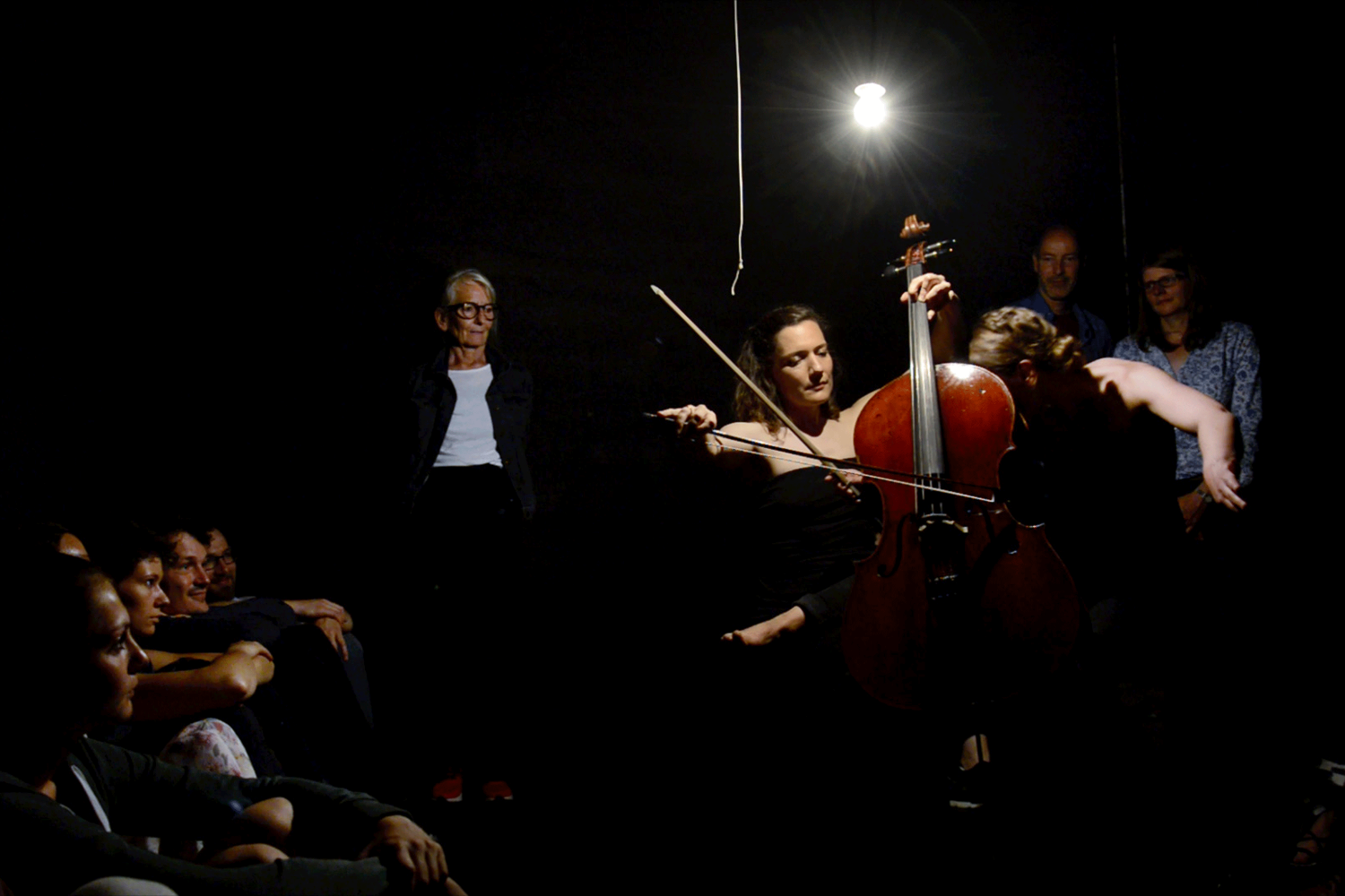 Eine Performerin am Cello ist in einem dunklen Raum zu sehen. Die Besucher*innen sitzen am Rand und schauen ihr zu.