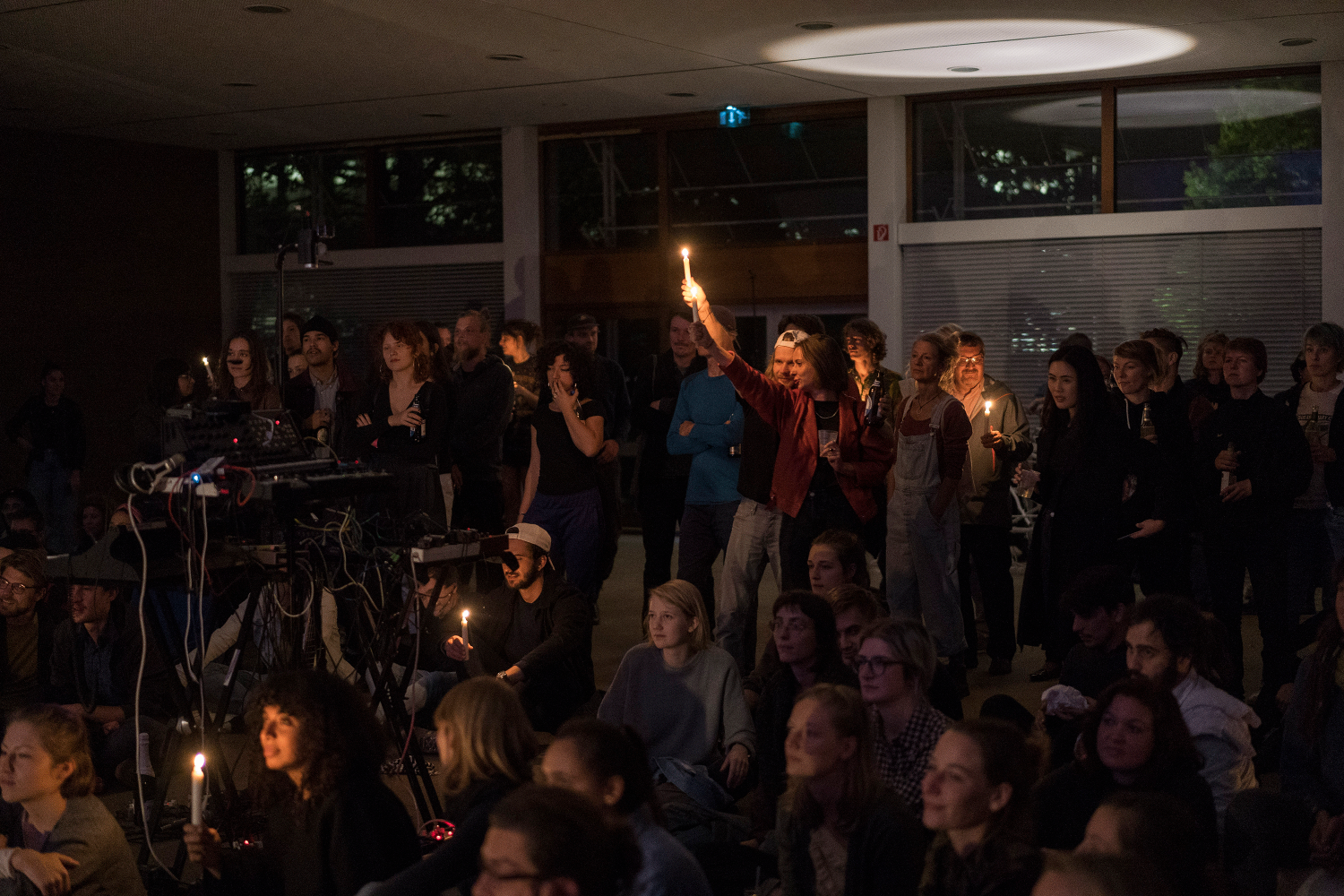 Das Publikum steht und sitzt bei der Performance von Contrau on Ice. Fünf Menschen haben Kerzen in der Hand. Draußen ist es dunkel.